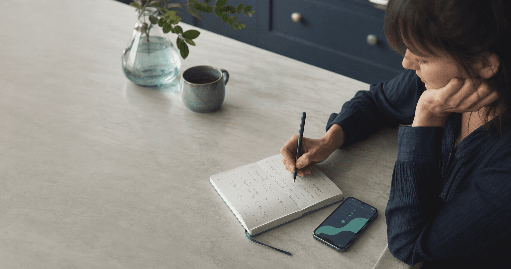Person writing headache symptoms in a notebook beside a phone