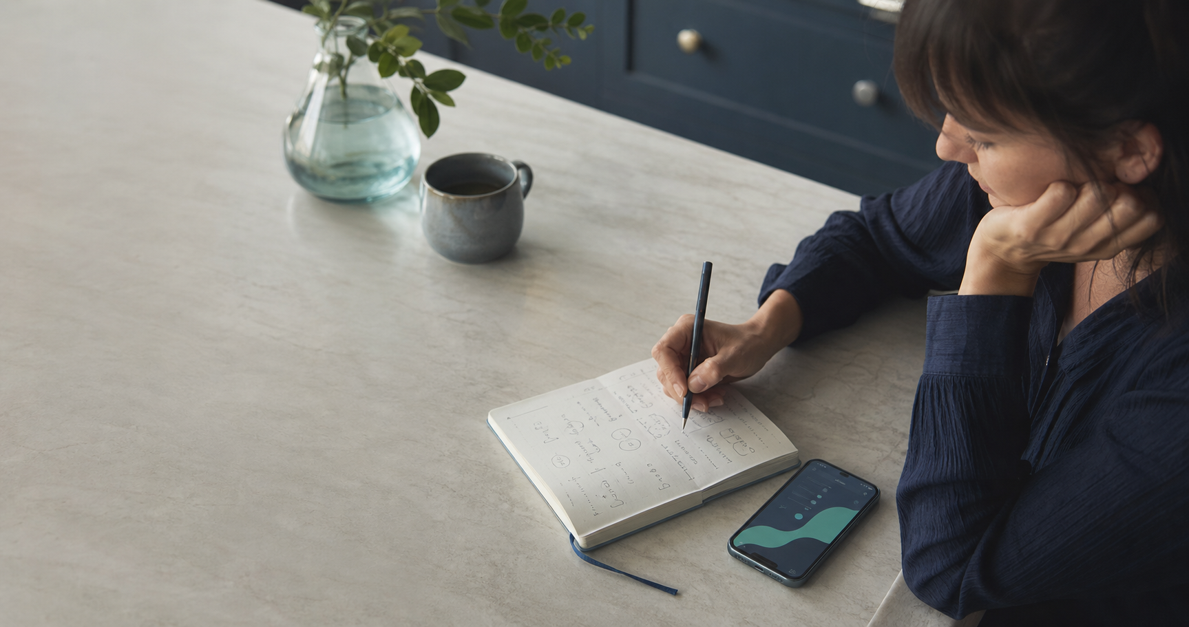 Person writing headache symptoms in a notebook beside a phone
